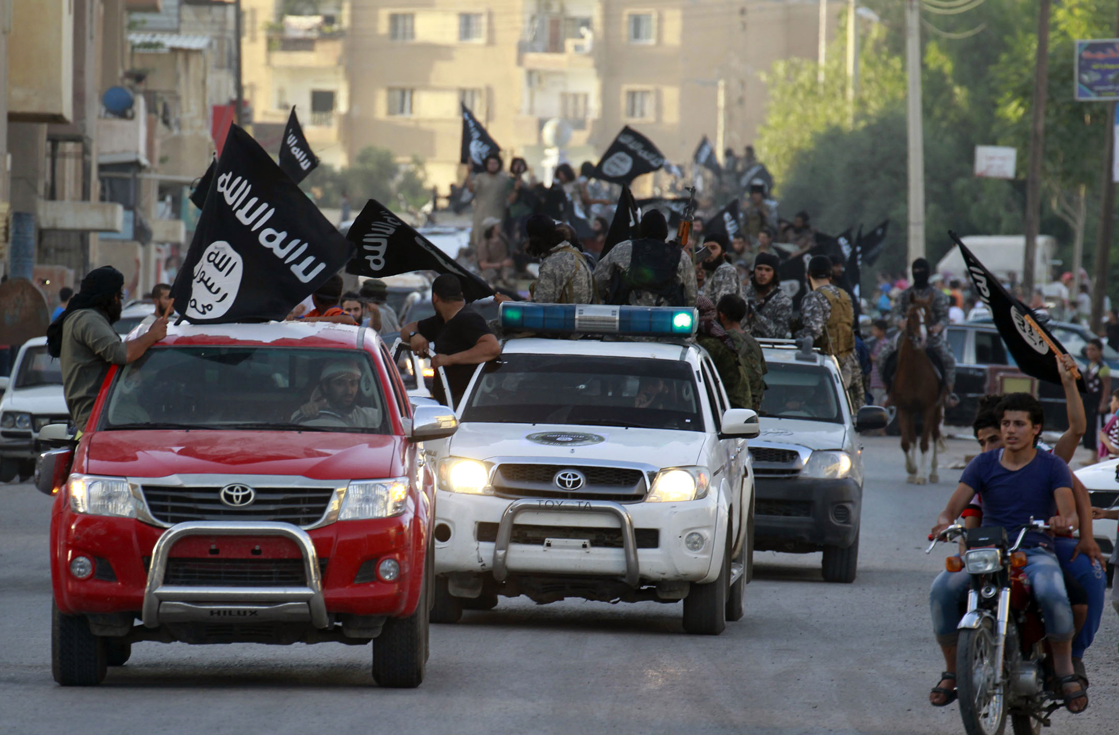 Luchadores militantes de Daesh ondeando banderas, viajan en vehículos, mientras participando en un desfile militar por las calles de la provincial norte Raqqa, Siria, 30 junio 2014