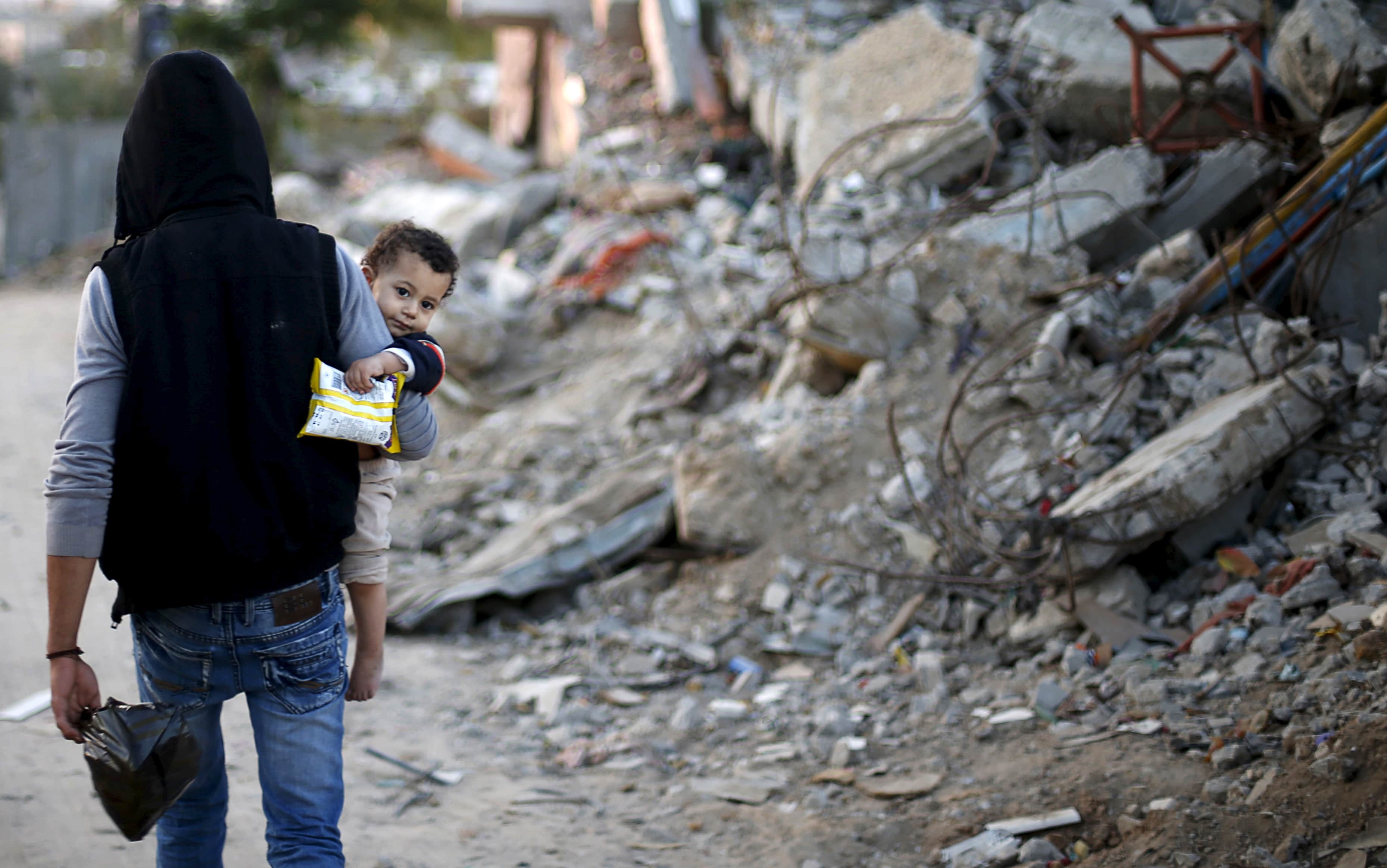 On 4 May 2015, a Palestinian man holding his son walks past houses in Gaza City destroyed during the previous summer's conflict, REUTERS/Mohammed Salem