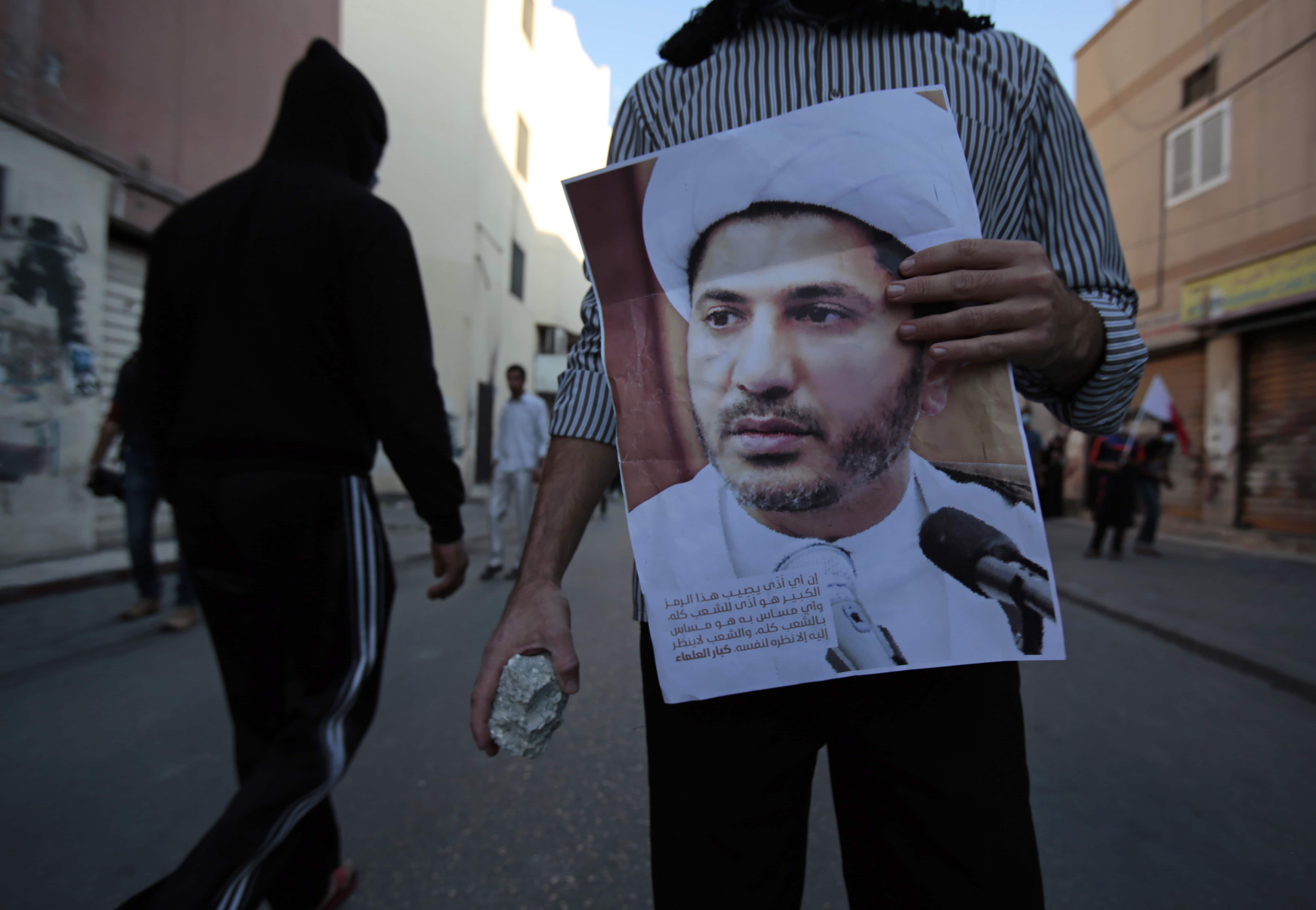 In this Saturday, Jan. 3, 2015 file photo, a Bahraini anti-government protester holding a stone and a picture of jailed Shiite cleric Sheik Ali Salman confronts police during clashes in Bilad Al Qadeem, Bahrain., AP Photo/Hasan Jamali, File