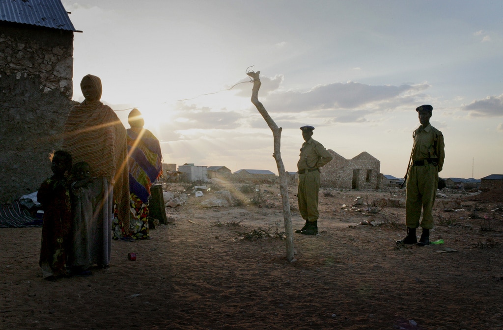 A woman and her children stand at a settlement for internally displaced persons in Somalia, with armed police standing by., Kate Holt/IRIN