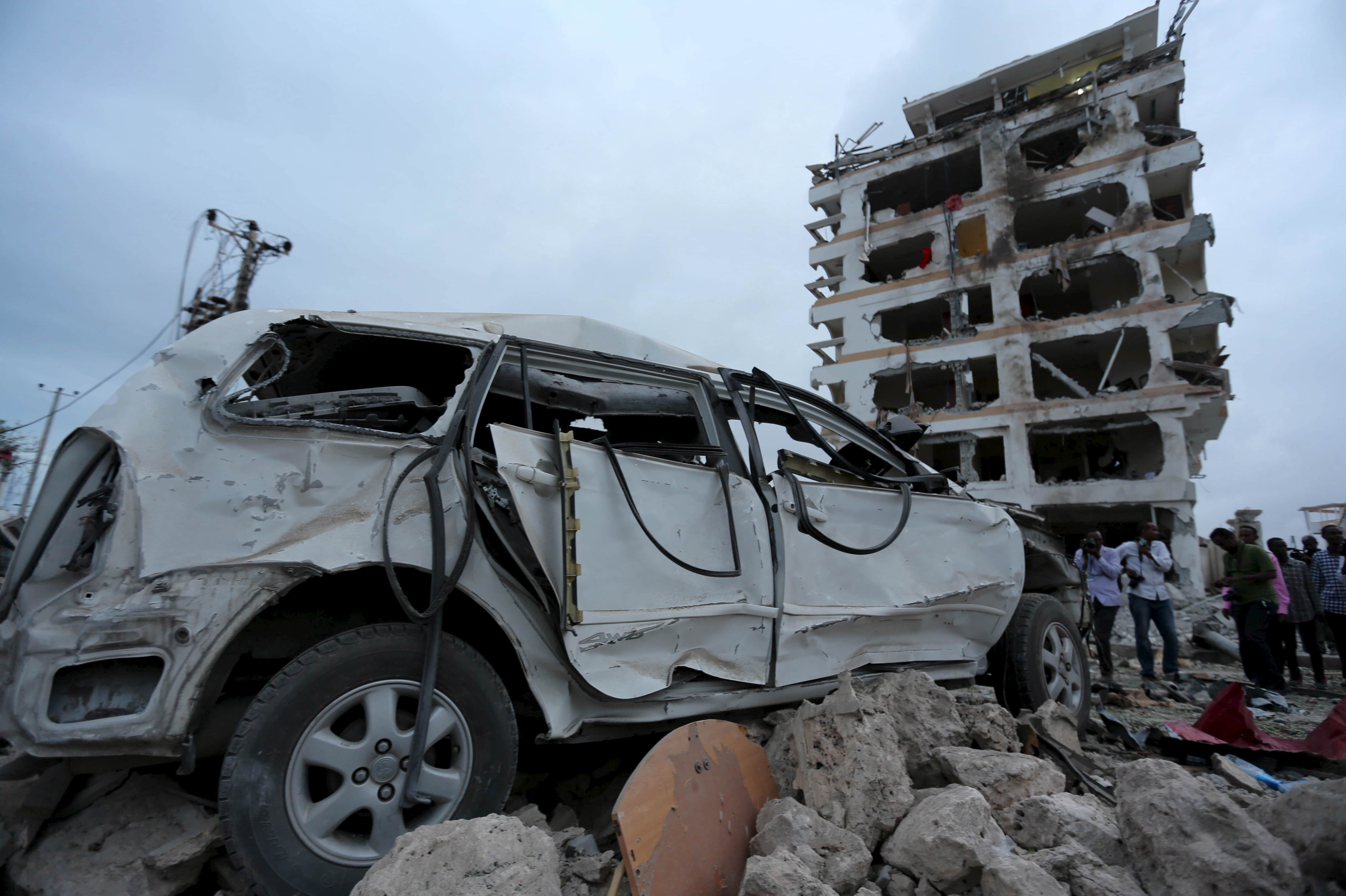 Somali government soldiers and journalists stand near a car destroyed in front of the Jazeera hotel after an attack in Mogadishu, 26 July 2015. , REUTERS/Feisal Omar