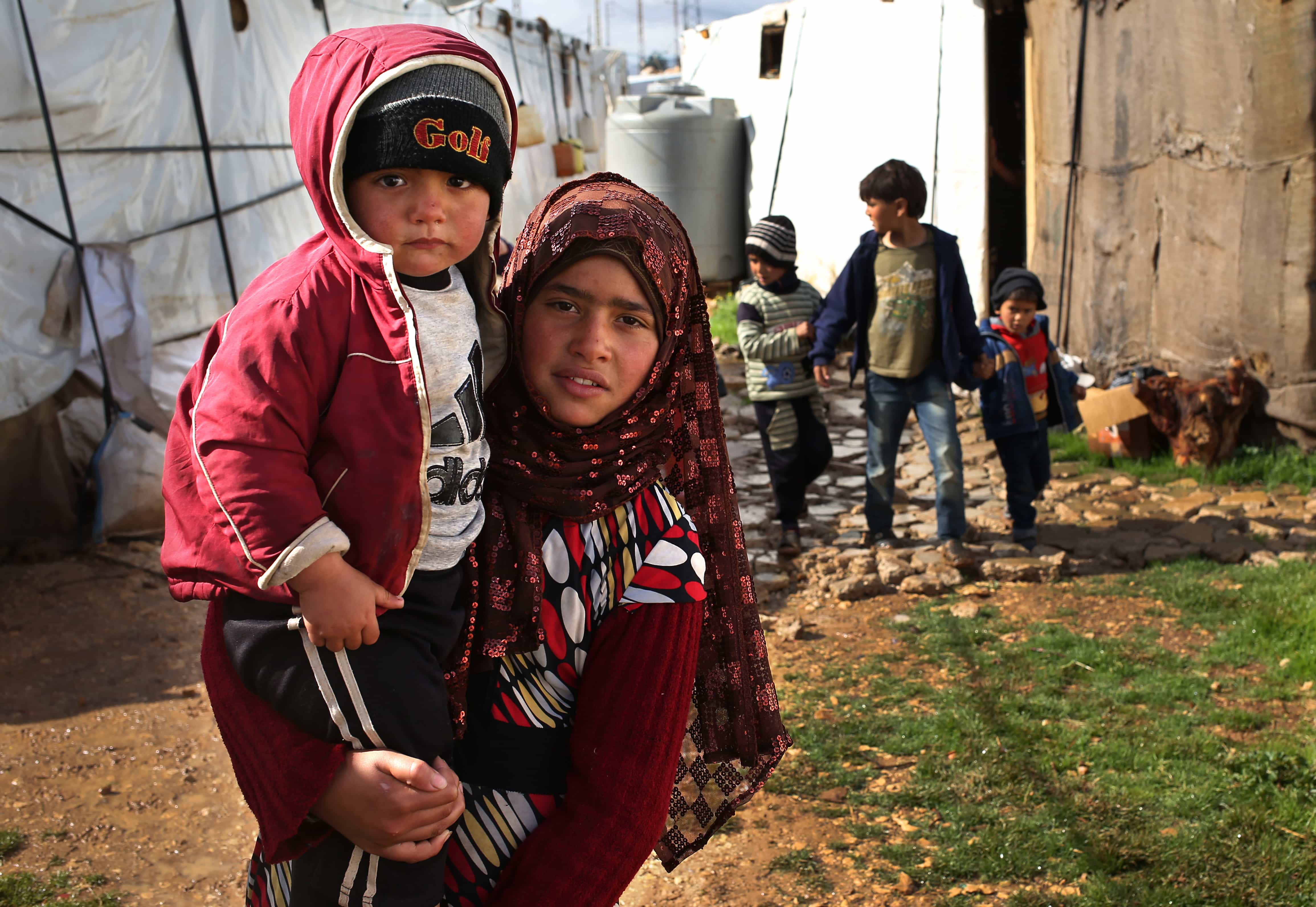 A Syrian girl carries her brother and walks between tents with other children at their refugee encampment in the Lebanese-Syrian border town of Majdal Anjar, AP Photo/Hussein Malla