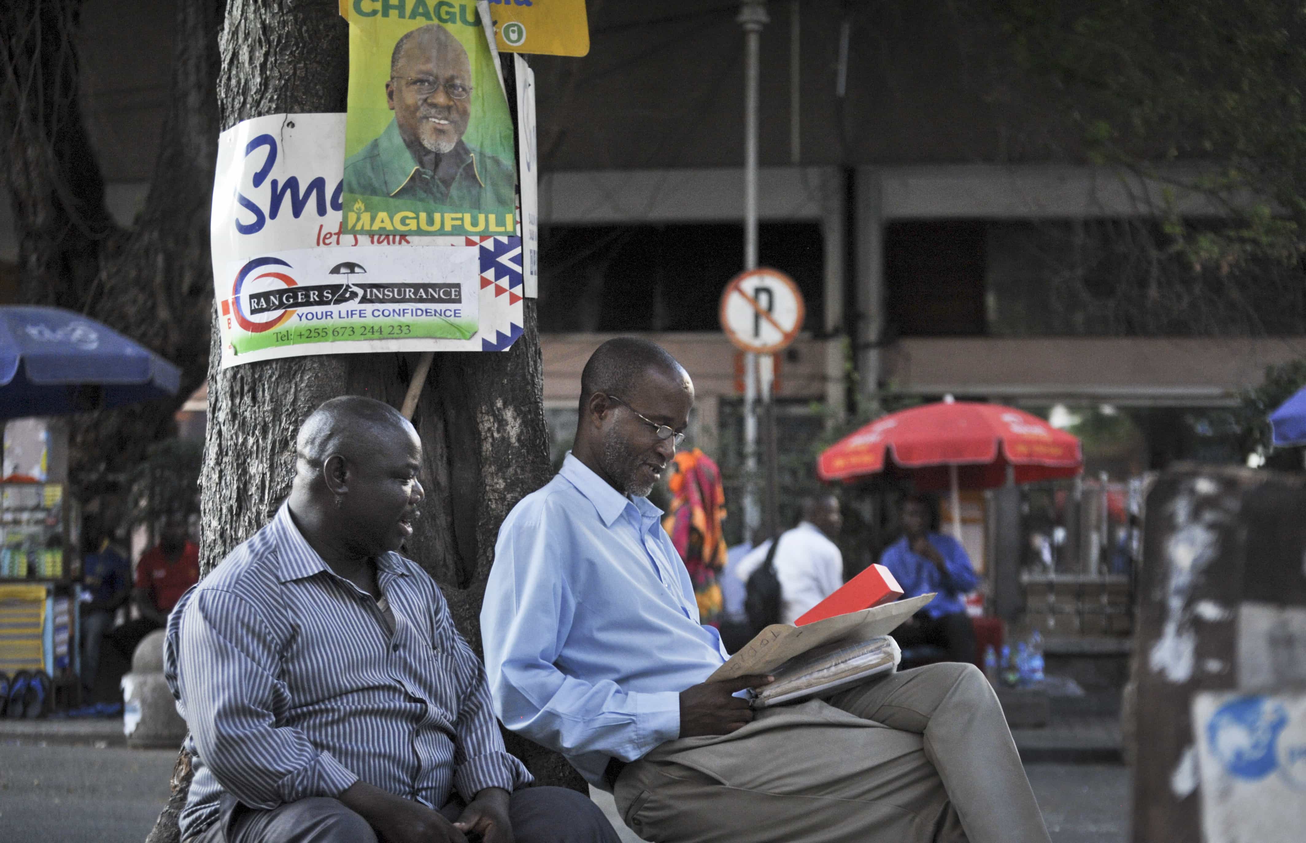 Tanzanians sit next to a tree, underneath an election poster for ruling party presidential candidate John Magufuli, as they await election results in Dar es Salaam, Tanzania 27 October 2015, AP Photo/Khalfan Said