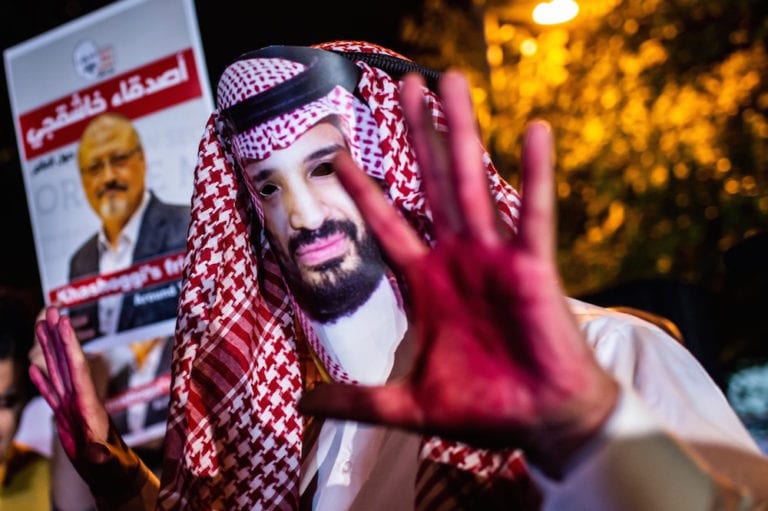 متظاهر يرتدي قناع لوجه ولي العهد السعودي الأمير محمد بن سلمان، ويداه باللون الأحمر بجانب أشخاص يحملون ملصقات للصحفي جمال خاشقجي، وذلك خلال مظاهرة خارج القنصلية السعودية في اسطنبول، تركيا، 25 تشرين الثاني 2018. (تصوير: ياسين أكغول / الوكالة الفرنسية عبر Getty Images).