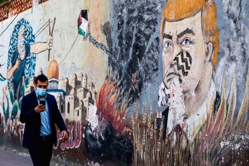 رجل يرتدي الكمامة بسبب فيروس كورونا المستجد، ويتصفح الهاتف وهو يمشي أمام كتابات تصور الرئيس الأمريكي المنتهية ولايته دونالد ترامب، في مدينة غزة، 8 تشرين الثاني 2020 . (الصورة من: محمد عابد / الوكالة الفرنسية عبر Getty Images).