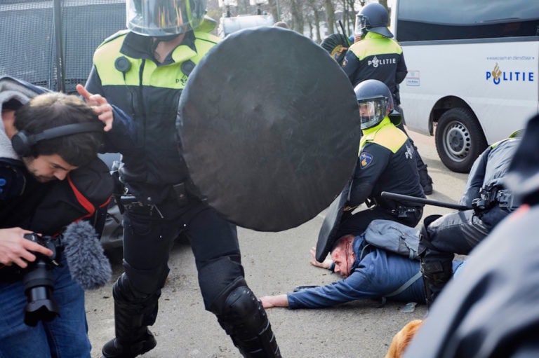 A journalist raises his arm to protect himself from a police officer as a man is seen bleeding in the background after being beaten by riot police, during a rally protesting COVID measures, The Hague, Netherlands, 14 March 2021, Pierre Crom/Getty Images