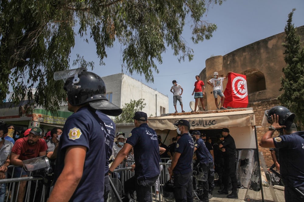 Supporters of President Kais Saied set up the Tunisian flag on the roof of a store as riot police stand guard in front of the parliament building, in Tunis, 26 July 2021, after Saied decided to freeze parliament and sack Hichem Mechichi’s government. Chedly Ben Ibrahim/NurPhoto