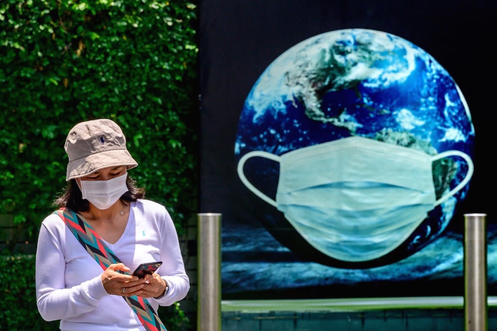 A woman checks her phone, while standing in front of a poster of the earth with a protective face mask during the COVID-19 pandemic, in Bangkok, Thailand, 22 April 2021, MLADEN ANTONOV/AFP via Getty Images